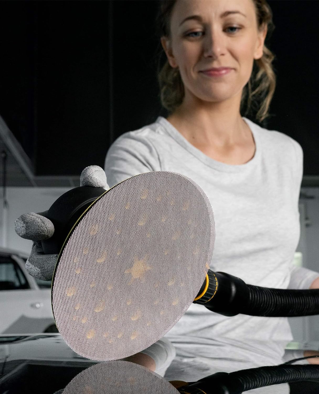 A woman holds a Mirka Abranet sanding disc while wearing protective gloves, showcasing the disc's mesh design for efficient dust extraction during sanding. She stands in a well-lit workshop setting.