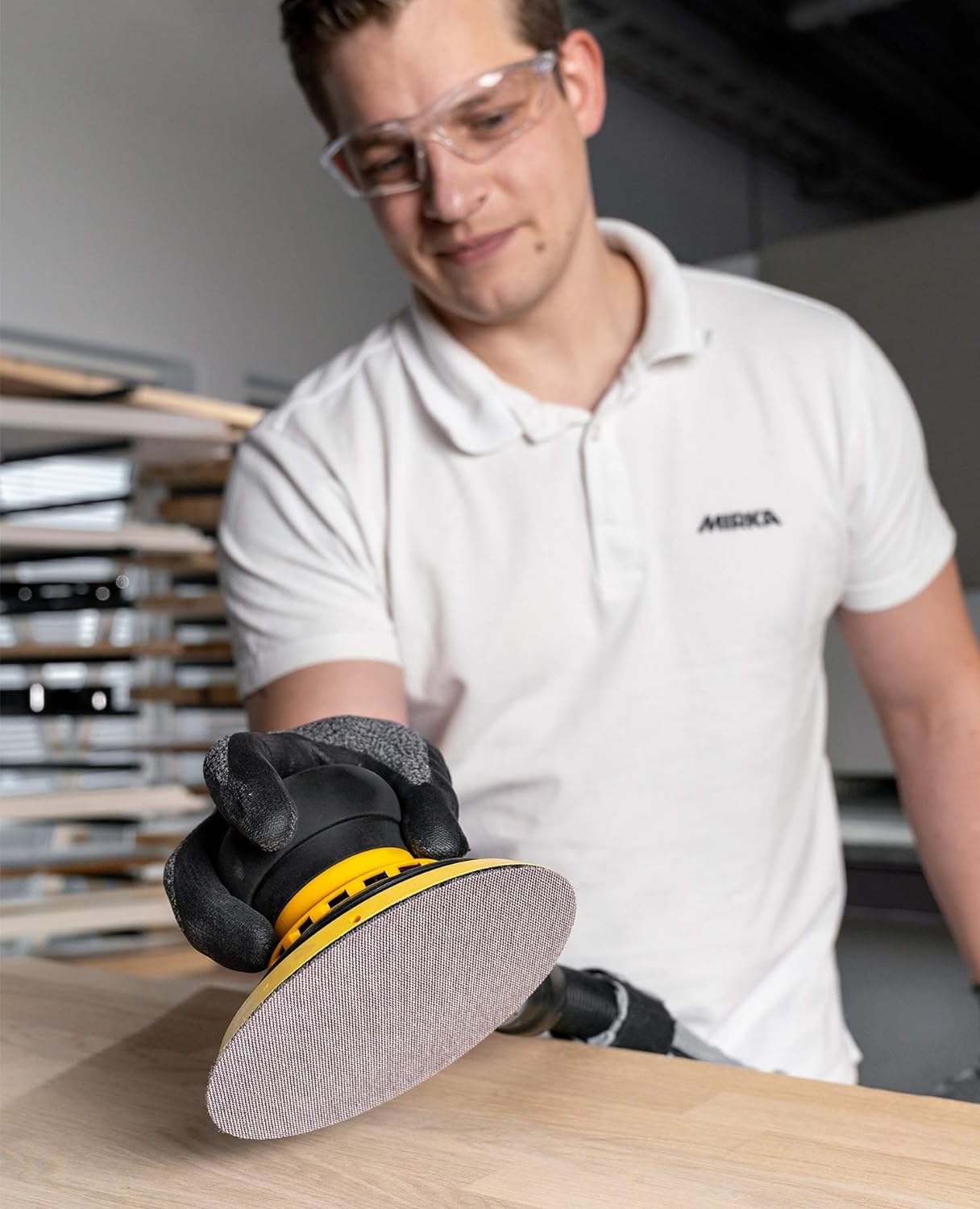 "A man wearing safety glasses and a white Mirka shirt demonstrates using a Mirka sanding tool with an Abranet disc on a wooden surface. He is holding the tool with gloved hands in a well-lit workshop environment.