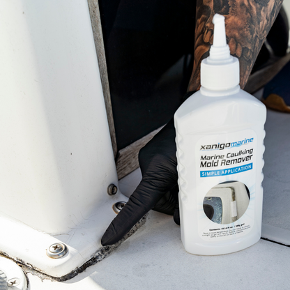 A hand wearing a black glove applying Xanigo Marine Caulking Mold Remover to a boat's caulking area, focusing on the removal of mold and mildew stains. The product is in the foreground with a close-up view of the surface being treated.