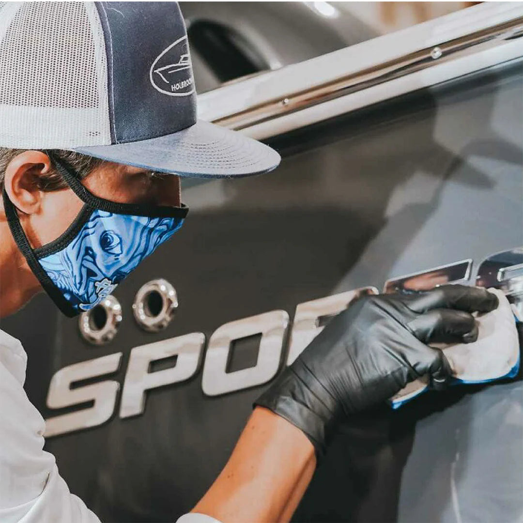 A person wearing a cap, mask, and gloves is polishing a boat's 'SPORT' decal using a cloth. The individual is focused on cleaning the boat’s surface near the decal, showcasing meticulous detailing.