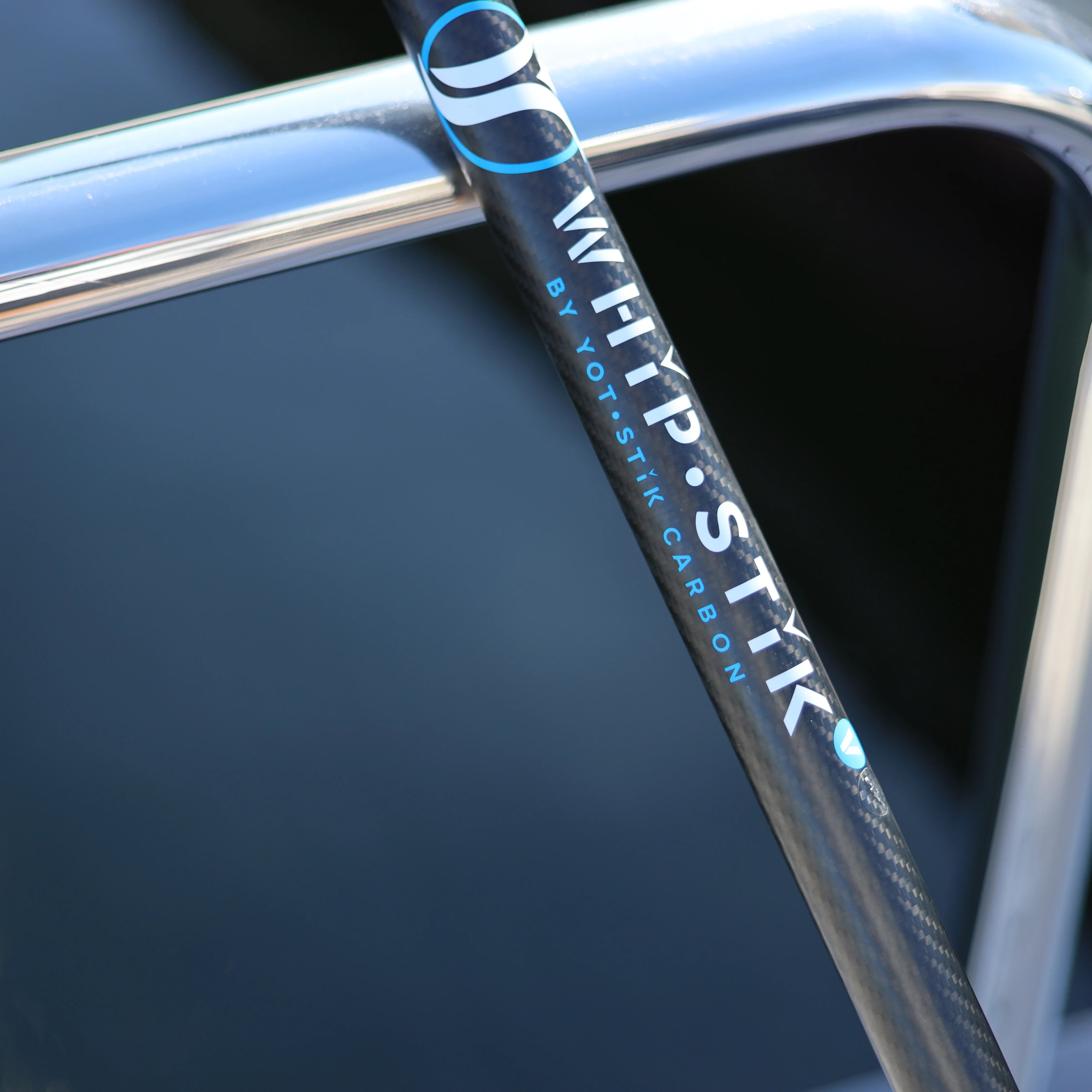 A close-up of a carbon fiber whip antenna with the brand name 'WHIP·STIK' displayed prominently. The antenna is mounted on a shiny, polished boat railing, showcasing its sleek design and high-quality craftsmanship.
