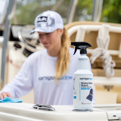 A woman wearing a hat and a long-sleeve shirt is cleaning a boat with a microfiber cloth. In the foreground, a bottle of Xanigo Marine Waterless Wash & Fabric Refresher is prominently displayed, showing its label and spray nozzle.