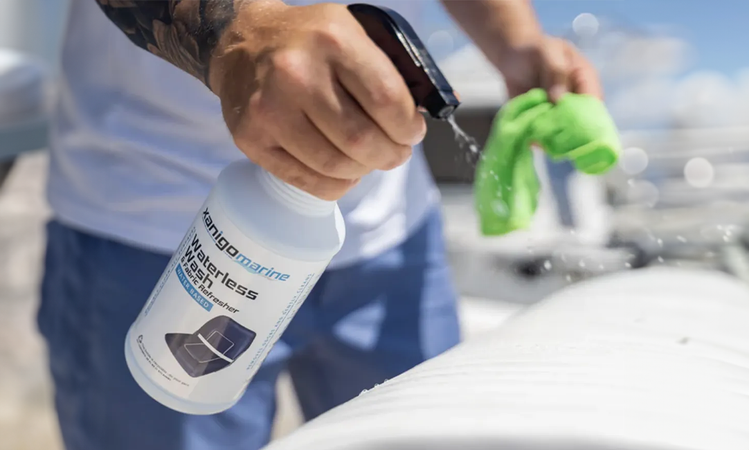 A person sprays Xanigo Marine Waterless Wash & Fabric Refresher onto a surface of a boat while holding a green microfiber cloth in their other hand, cleaning the surface in the bright sunlight.