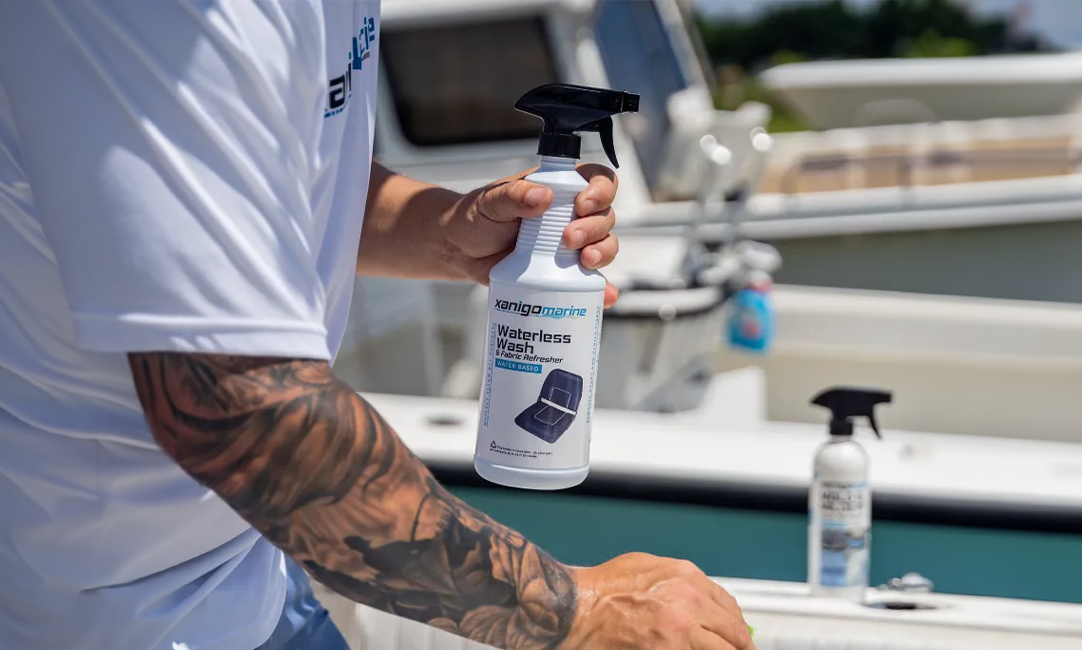 A person with tattoos on their arm is holding a bottle of Xanigo Marine Waterless Wash & Fabric Refresher in one hand, ready to spray, with a bottle of Xanigo Marine Mold & Mildew Stain Remover visible in the background on a boat.