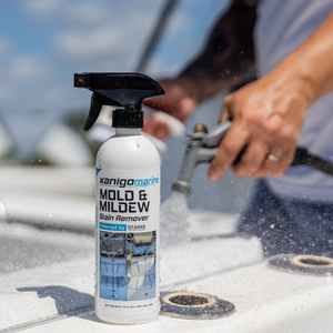 Close-up of a Xanigo Marine Mold & Mildew Stain Remover spray bottle on a boat's surface, with a person in the background using a hose to clean the area. The bottle prominently features the 'Powered by STARKE' label.