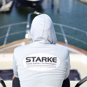 A person wearing a light-colored hoodie with the 'STARK' logo on the back, sitting on the deck of a yacht, overlooking the water and docks.