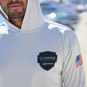 A person wearing a white hoodie with the 'STARK' logo and 'Certified Installer' badge on the chest, along with an American flag on the sleeve, smiling slightly. The background shows a blurred marina with boats.